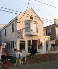 several people are walking in front of a white building on a street corner with two story houses behind them