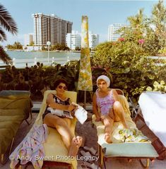 two women sitting on lounge chairs in front of a cityscape with palm trees