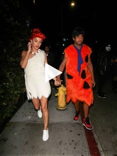 two people in costumes walking down a sidewalk at night with fire hydrant behind them