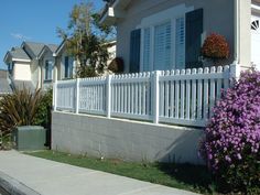 a white picket fence in front of a house with purple flowers growing on the side