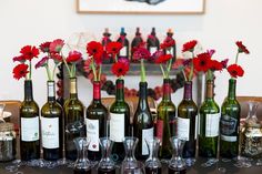 a table topped with lots of bottles of wine and vases filled with red flowers