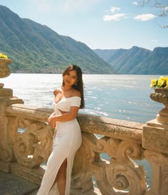 a woman in a white dress standing on a balcony next to the water and mountains