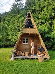 a woman standing in front of a small wooden cabin with logs on the ground and trees behind her