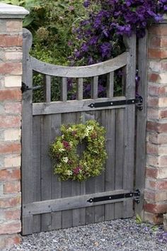 a wooden gate with a wreath hanging on it's side, surrounded by flowers