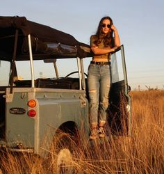 a woman standing next to an old jeep in the middle of a field with her hand on her head