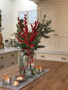 a vase filled with red berries and greenery on top of a wooden table next to candles