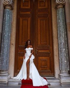 a woman in a white dress is standing on a red carpet near a door with columns