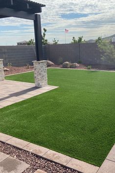 an outdoor area with artificial grass and stone pillars in the foreground, surrounded by landscaping