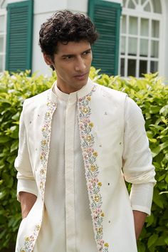 a man wearing a white suit and jacket with flowers on the lapel, standing in front of bushes