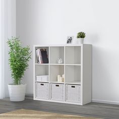 a white bookcase with baskets on the bottom and two plants in front of it