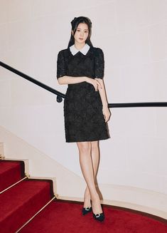 a woman in black dress standing on red carpeted stairs with her arms crossed and looking at the camera