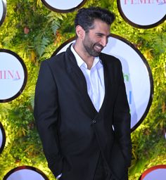 a man in a black suit and white shirt standing next to a wall with plants on it