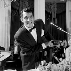 a man in a tuxedo standing next to a microphone at a dinner table