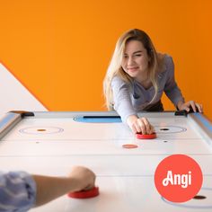 two people playing air hockey with an orange wall behind them and an angi logo on the front