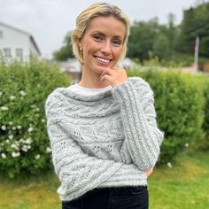 a woman standing in front of some bushes wearing a gray sweater and black pants with her hand on her chin