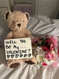 a teddy bear sitting on top of a bed next to a bouquet of flowers and a sign that says will you be my valentine?