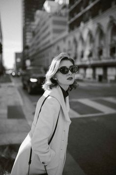 black and white photograph of woman in sunglasses on street corner with buildings behind her, looking off to the side