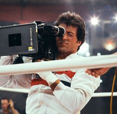 a man holding a camera in front of his face while standing next to a rope