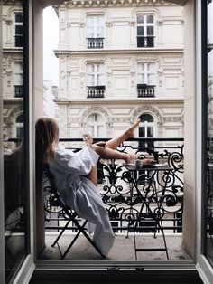 a woman sitting in a chair on top of a balcony next to a building and looking out the window