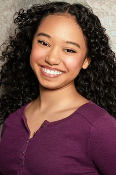 a woman with long curly hair smiling at the camera while wearing a purple shirt and standing in front of a wall