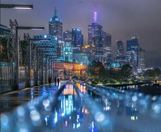 the city skyline is lit up at night with lights reflecting in the water and people walking on the sidewalk