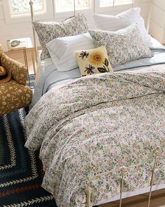 a bed with floral comforter and pillows in a room next to a chair, rug and window