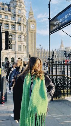 a woman standing on the sidewalk with a green scarf around her neck and a clock tower in the background