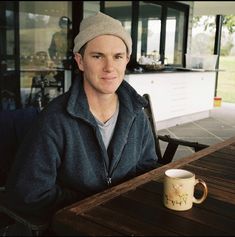 a man sitting at a table with a coffee cup in front of him and looking into the camera