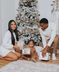 a man and woman opening presents next to a christmas tree with a small child sitting on the floor