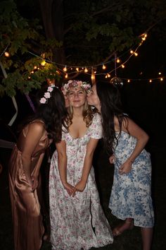 three girls in dresses standing next to each other with lights strung over the trees behind them