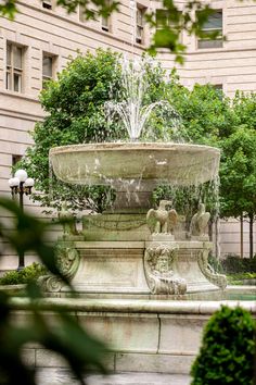 a water fountain surrounded by trees in front of a large beige building with many windows