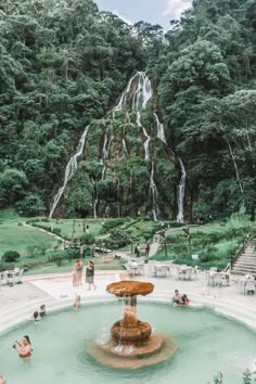 people are swimming in the water near a fountain with a waterfall on top and trees surrounding it