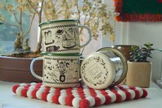 three coffee mugs sitting on top of a red and white striped mat next to a potted plant