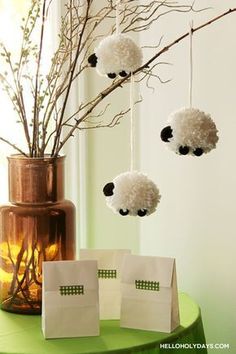 three small white sheep hanging from a branch in front of a vase filled with flowers