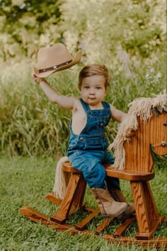 a little boy sitting on top of a wooden rocking horse with a hat in his hand