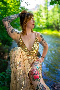 a woman with tattoos sitting on the edge of a river wearing a gold dress and holding her hair up