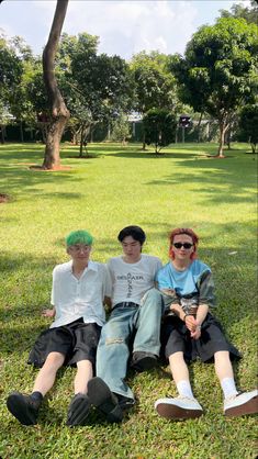 three young men sitting on the grass in front of some trees and one is wearing green hair