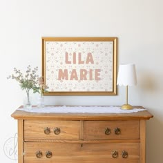 a wooden dresser with flowers on top and a framed poster above it that says lila marie