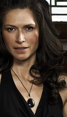 a woman with long dark hair wearing a black top and necklace, looking at the camera