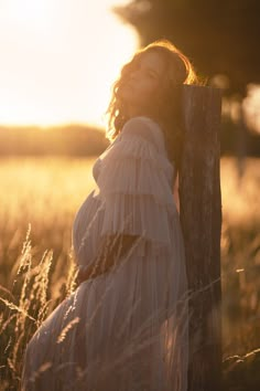 a pregnant woman leaning against a wooden post in a field at sundown with her eyes closed