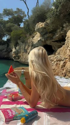 a woman laying on top of a beach next to the ocean holding a slice of watermelon