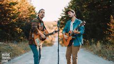 two men are playing guitars and singing into microphones while standing on a road with trees in the background