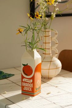 an orange juice carton sitting on top of a table with flowers in the vase