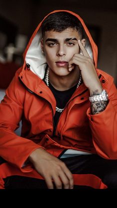 a young man in an orange jacket sitting on a bed with his hand to his face