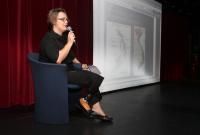 a woman sitting in a chair talking on a stage with a red curtain behind her