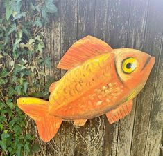 an orange fish with yellow eyes is hanging on a wooden fence next to some green plants