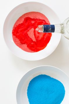 two bowls with red and blue powder in them next to a bottle of water on a white surface