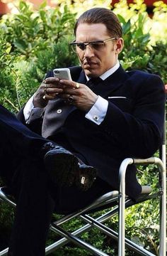 a man sitting in a chair looking at his cell phone while wearing glasses and a suit