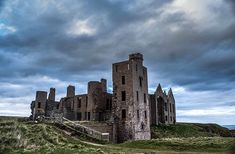 an old castle sitting on top of a lush green hillside under a cloudy sky with dark clouds