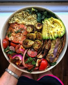 a person holding a large bowl filled with different types of vegetables and meat on top of rice